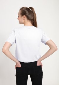 Camisa de algodón blanca con mangas cortas, cuello redondo y corte relajado, vista desde la parte de atrás. Llevada con vaqueros negros que tienen bolsillos traseros.