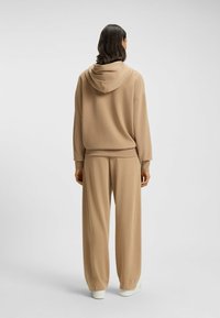 Set di felpa beige e pantaloni a gamba larga realizzati in tessuto morbido, con cappuccio, vestibilità rilassata e polsini a coste. Le scarpe sono bianche.