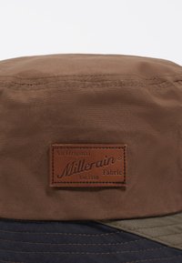 Sombrero tipo bucket de tela marrón con una etiqueta de cuero Millerain, que presenta un ala cosida y una textura suave. Incluye acentos en azul marino oscuro y verde oliva.