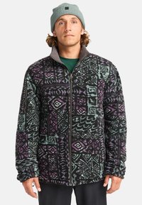 Chaqueta de forro polar con base negra, que presenta un patrón geométrico multicolor en púrpura y verde azulado. Cierre de cremallera, cuello alto y mangas largas.