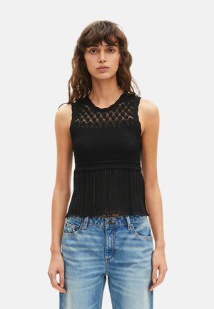 Vrouw met golvend bruin haar, gekleed in een mouwloos zwart gebreid topje met patronen en lichtblauwe spijkerbroek, staand tegen een effen achtergrond.