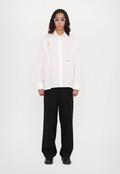 Chemise blanche à manches longues avec un col boutonné et une coupe décontractée, arborant un petit logo bleu. Assortie à un pantalon noir à jambe large et des chaussures.