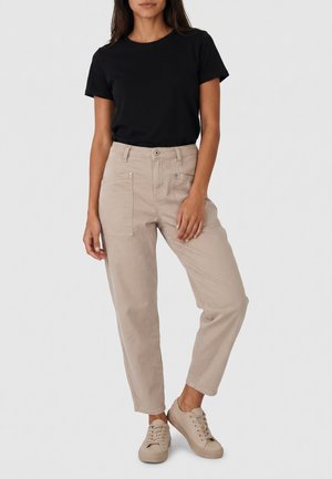 Femme portant un t-shirt noir à manches courtes, un pantalon beige taille haute avec poches avant, et des baskets beiges, debout devant un fond uni.