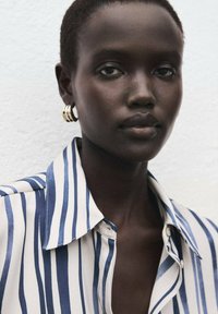 Črtasta srajca v kremni in mornarsko modri barvi, gladka tkanina s ovratnikom. Poudarjeni zlati obroči za ušesa, posnetek obraza od blizu.