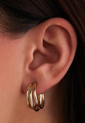 Goldene Creolen mit einem wellenförmigen Design, die aus drei miteinander verbundenen Ringen bestehen. Glatte Oberfläche mit einer reflektierenden Oberfläche.