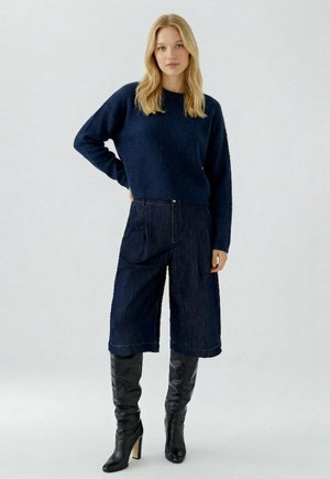 Femme debout portant un pull bleu marine, un jean large court foncé, et des bottes noires montantes à talons hauts sur un fond uni.