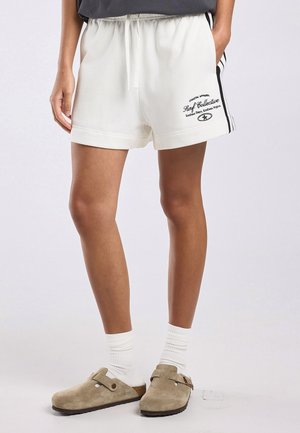 Persoon die witte shorts met trekkoord draagt met zwarte zijstrepen en logo, witte sokken en beige Birkenstock klompen, staand tegen een effen achtergrond.