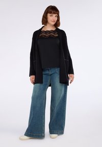 Donna che indossa un top nero con dettagli in pizzo, un cardigan nero, jeans blu a gamba larga e scarpe bianche, in piedi contro uno sfondo semplice.