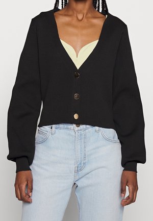 Žena v černém krátkém kardiganu se třemi kovovými knoflíky přes světle žlutý top, v kombinaci s modrými džíny s vysokým pasem.