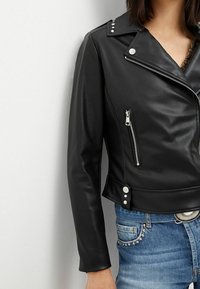 Giacca da moto in pelle nera con borchie argentate sulle spalle, chiusura con zip sul davanti e tasche, texture liscia, design aderente.