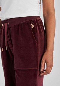 Vínové tepláky z velurového materiálu s elastickým pasem, stahovacím šňůrkem, bočními kapsami a zlatým logem na horní levé stehně.