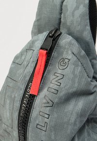 Grått stoffryggsekk med et teksturert mønster, med en fremtredende rød glidelåstrekk og svart glidelås, og ordet "LIVING" trykt på siden.