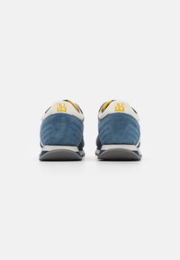 Sneaker in suede blu con un logo bianco e giallo sul tallone, dotate di una suola imbottita e pannelli laterali texturizzati per il supporto.