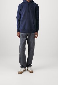 Sudadera azul marino con un bolsillo frontal y capucha con cordón, combinada con jeans gris claro y zapatillas blancas con detalles en marrón.