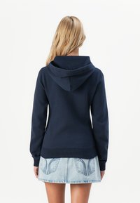 Woman with long blonde hair wearing a navy hoodie and light blue denim skirt, standing with her back to the camera on a white background.