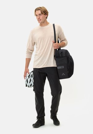 Sac à dos noir avec plusieurs compartiments, sangle en tissu. Le modèle porte une chemise beige à manches longues et un pantalon noir, en tenant un casque blanc.