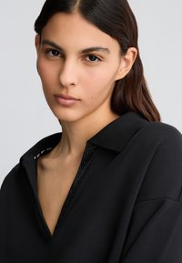 Vêtement noir avec un col, présentant un détail texturé et de perles au niveau de l'encolure. Matière douce, coupe décontractée, et couture visible le long du bord du col.