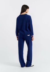 Middengrijze loungewear-set van zachte stof, bestaande uit een sweater met ronde hals en gestreepte manchetten, en bijpassende broek met een logo-patch op de achterkant.