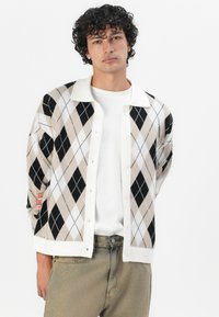 Argyle knit cardigan in black, beige, and white, featuring a white collar and button front, worn over a white shirt and beige trousers.