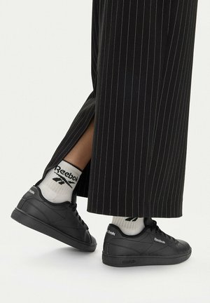 Černé tenisky Reebok s bílými ponožkami s logem, nošené s černými širokými kalhotami s pruhy a rozparkem u kotníku.