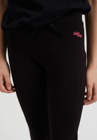 Črne legice iz mehkega materiala, z elastičnim pasom in vrvico. Vidna je majhna roza oznaka "Uživaj danes".