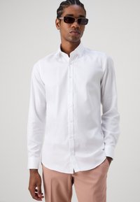 Chemise blanche à manches longues en tissu lisse, avec un devant à boutons et un ourlet arrondi. Associée à un pantalon rose clair.