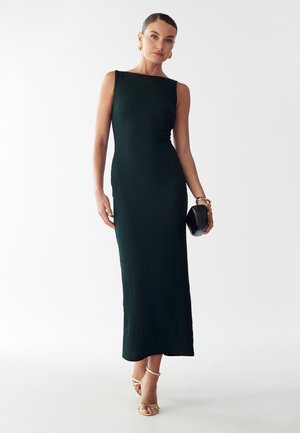 Žena v černých texturovaných maxi šatech bez rukávů, se zlatými kruhovými náušnicemi, náramkem, držící černý psaníčko, v béžových sandálech na vysokém podpatku, pózující.