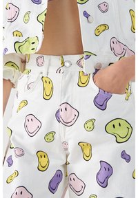 Conjunto de denim blanco con estampados de caras sonrientes coloridos en rosa, amarillo, verde y púrpura. Incluye detalles de botones y bolsillos frontales.