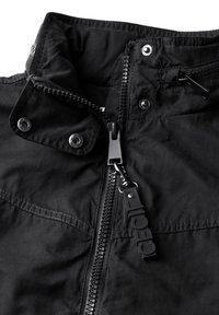 Black jacket with a high collar, featuring a front zipper and snap buttons. Made of textured fabric, includes a branded zipper pull and side pocket.