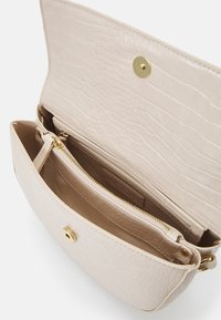 Krémová texturovaná kožená crossbody taška se zlatým magnetickým uzávěrem a vnitřní kapsou na zip, otevřená a ukazující detaily interiéru.