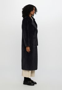 Long, dark navy coat made of plush fabric with a double-breasted design, large collar, and black buttons, paired with beige trousers.