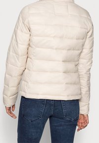 Světlobéžová péřová bunda s horizontálním prošíváním, stojáčkem a elastickými manžetami, kombinovaná s tmavými džíny.