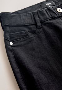 Close-up van de taille van een zwarte spijkerbroek met zakken, riemlussen, zilveren knoop en "NEXT" label op de binnenste tailleband.