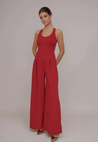 Combinaison sans manches rouge avec un corsage ajusté, des jambes larges et des détails plissés. Tissu lisse de couleur unie. Chaussures dorées.
