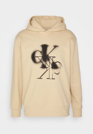 Haut à capuche beige clair avec une coupe ample, présentant un design graphique noir de lettres stylisées à l'avant et une capuche avec cordon de serrage.