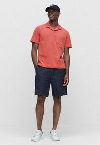Koraalrood poloshirt met korte mouwen en een borstzak, gecombineerd met navy shorts. Schoenen zijn witte sneakers met groene accenten.
