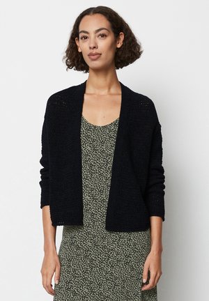 Black knitted cardigan with long sleeves and open front, worn over a patterned green dress. Textured fabric and draped design.