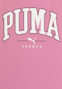 Camiseta rosa con "PUMA" en letras blancas de gran tamaño, con un contorno gris; incluye el texto "SPORTS" y un pequeño logo de puma.