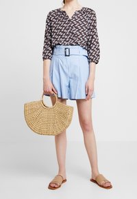 Blusa marrone a fantasia, pantaloni corti in denim azzurro chiaro a vita alta con cintura, borsa in paglia intrecciata e sandali piatti color nocciola.