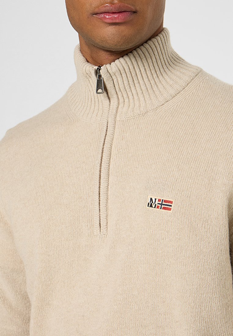 Pull en beige avec col zippé, fabriqué en tissu doux. Il est orné d'un petit logo brodé avec des accents rouges et bleus.