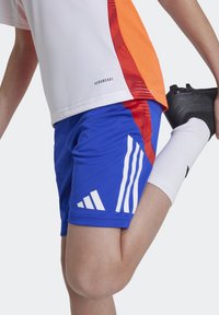 Siniset urheilushortsit valkoisilla Adidas-raidoilla, joissa on AeroReady-merkintä. Materiaali vaikuttaa sileältä ja kevyeltä, sopiva urheilutoimintaan.