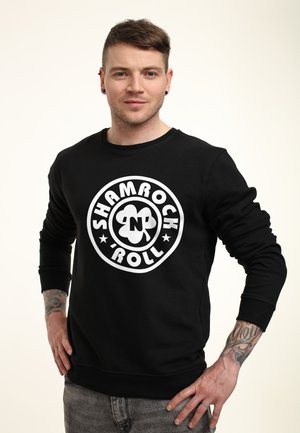 Mężczyzna z krótkimi włosami i kolczykami w uszach, ubraną czarną bluzę z kapturem z logo "Shamrock N Roll" i widocznymi tatuażami na obu przedramionach.