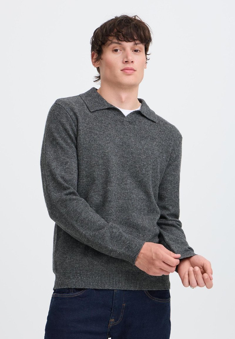 Jeune homme ajustant le poignet d'un pull en maille gris foncé avec col, portant un jean bleu foncé, sur un fond uni clair.