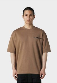Brun oversized bomulds T-shirt med ribbet halsudskæring, korte ærmer og sorte tekstgrafikker på forsiden. Sorte bukser nedenfor.