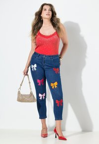 Červený top bez rukávů s krajkovým lemováním a ozdobami, modré džínové kalhoty s barevnými aplikacemi mašlí a třpytivá kabelka. Červené vysoké podpatky.