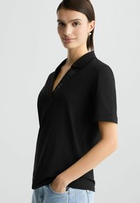 Zwarte polo in zachte stof, met een V-hals en decoratieve accenten, korte opgerolde mouwen en een relaxte pasvorm.