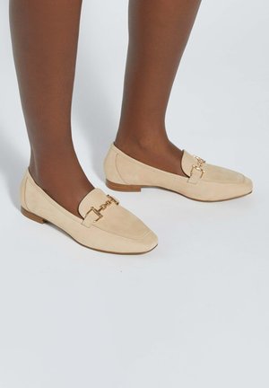 Pieds à la peau brune portant des mocassins en daim beige avec un détail en métal doré en forme de mors et de petits talons en bois, sur un fond blanc.
