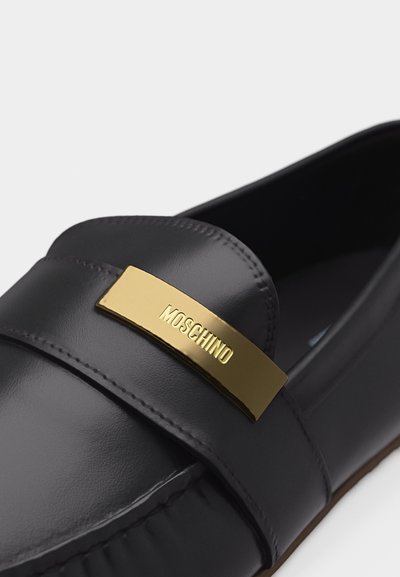 Gros plan d'un mocassin en cuir noir avec une plaque en métal doré gravée "MOSCHINO" sur la bride à l'avant.