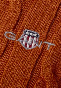 Oranges Strickstoff mit geripptem Muster, das ein kleines gesticktes Wappen mit Sternen und Streifen sowie teilweise Text in grauem Faden zeigt.