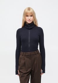 Maglione dolcevita a coste blu navy con zip frontale completa, maniche lunghe e vestibilità aderente, abbinato a pantaloni marrone chiaro a gamba larga.